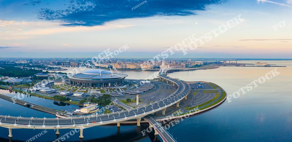 Фотография панорамного вида Санкт-Петербург, Россия, сделанная с высоты птичьего полёта в период белых ночей. На изображении показаны воды Нева и Финский залив, а также Крестовский остров с современной скоростной автомагистралью. Фотоснимок передаёт масштаб города, его прибрежные пространства и сочетание природного ландшафта с развитой транспортной инфраструктурой. Фото отражает атмосферу белых ночей и уникальный облик Санкт-Петербурга как крупного северного мегаполиса. Иллюстрация может использоваться как изображение, картинка или слайд, посвящённые панорамам и городам России.